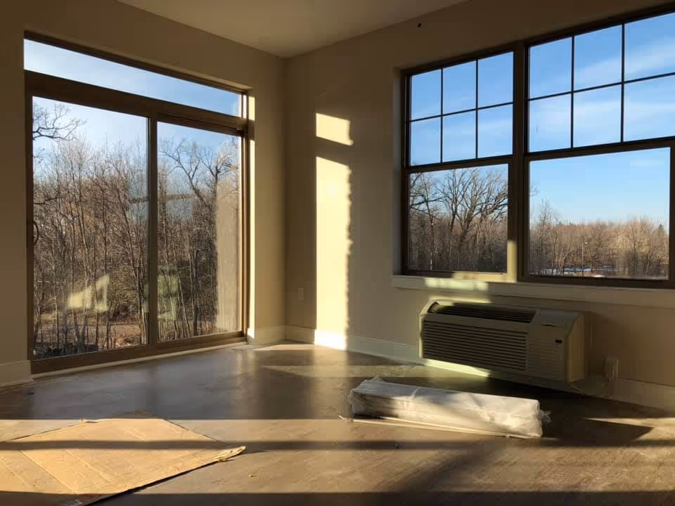 Empty room with large windows showing a view of leafless trees outside. The room has a floor-mounted air conditioning unit below one window and some cardboard and a wrapped item on the floor. Sunlight casts shadows on the walls and floor.