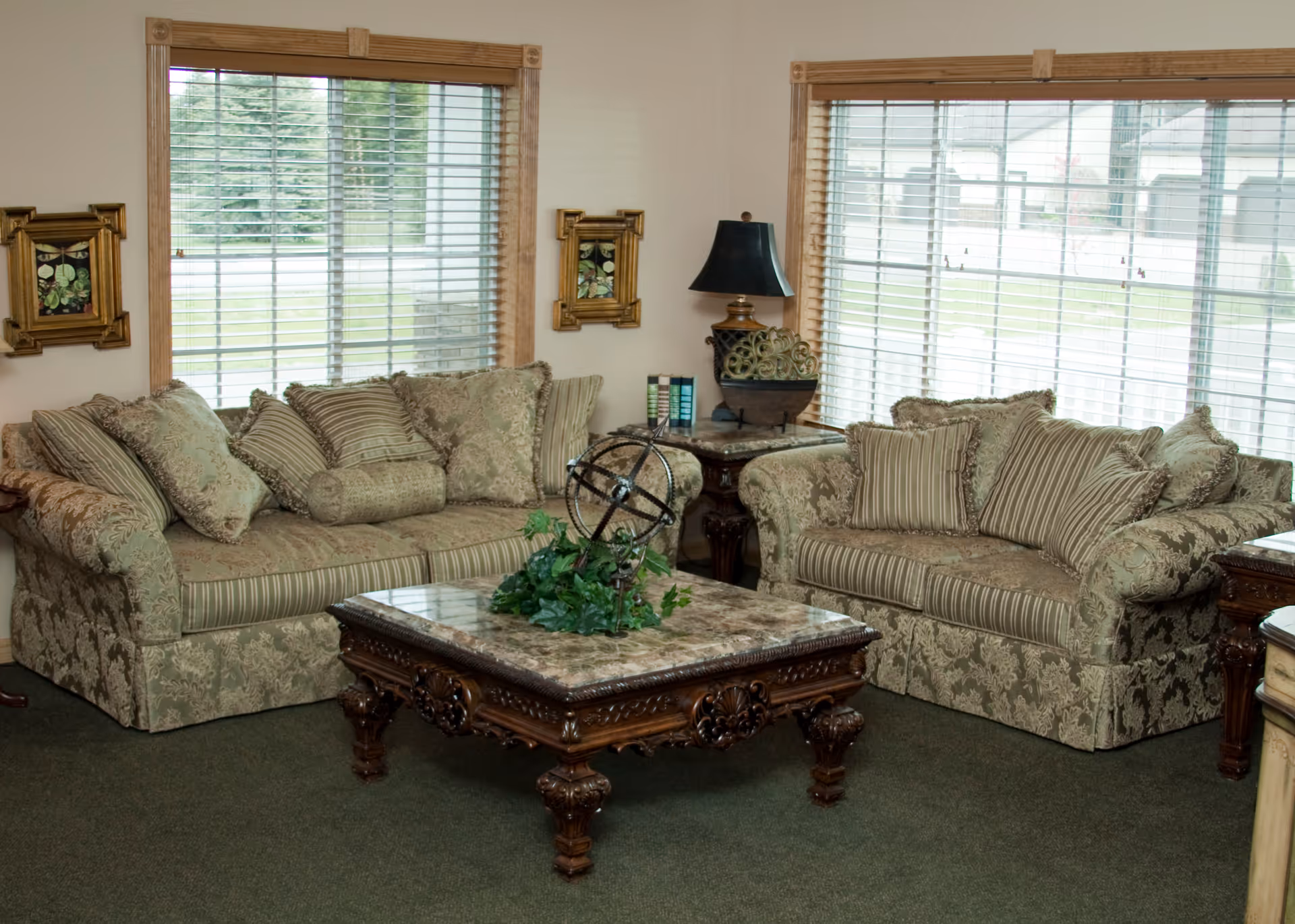 A cozy living room with two patterned sofas adorned with multiple cushions, a decorative wooden coffee table with a marble top and a green plant centerpiece, a side table with a lamp and books, and large windows with wooden blinds letting in natural light.