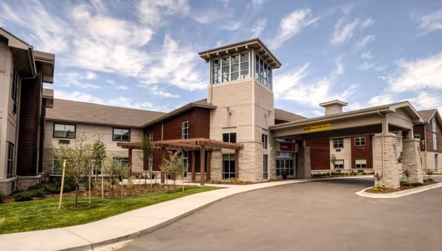 Front exterior of a senior living facility showing the entrance drive, porte-cochere, landscaping, and a multi-story tower.