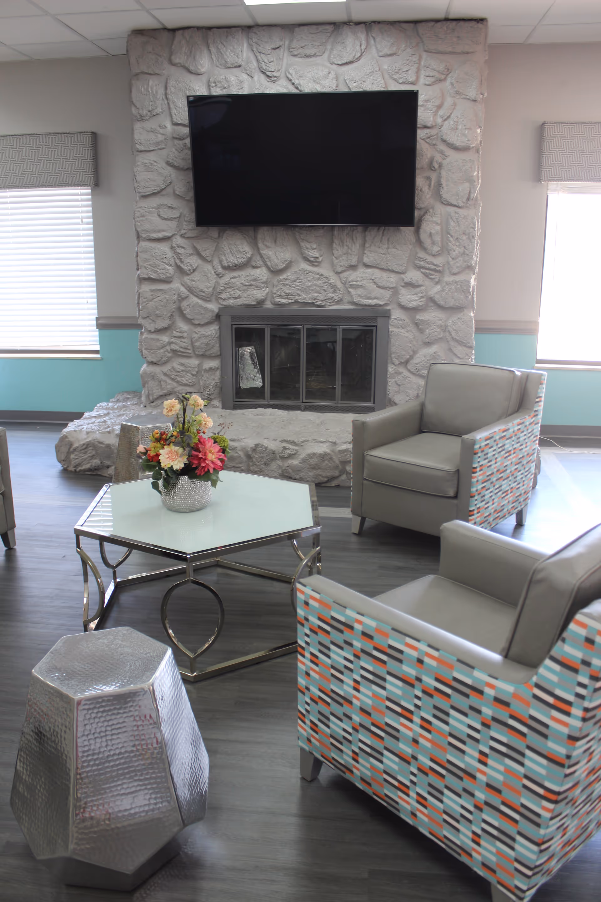 A cozy sitting area with two modern armchairs featuring a colorful geometric pattern on the sides, a hexagonal glass-top coffee table with a flower arrangement, a metallic geometric stool, and a stone fireplace with a mounted flat-screen TV above it. The room has large windows with blinds and light blue walls with gray trim.
