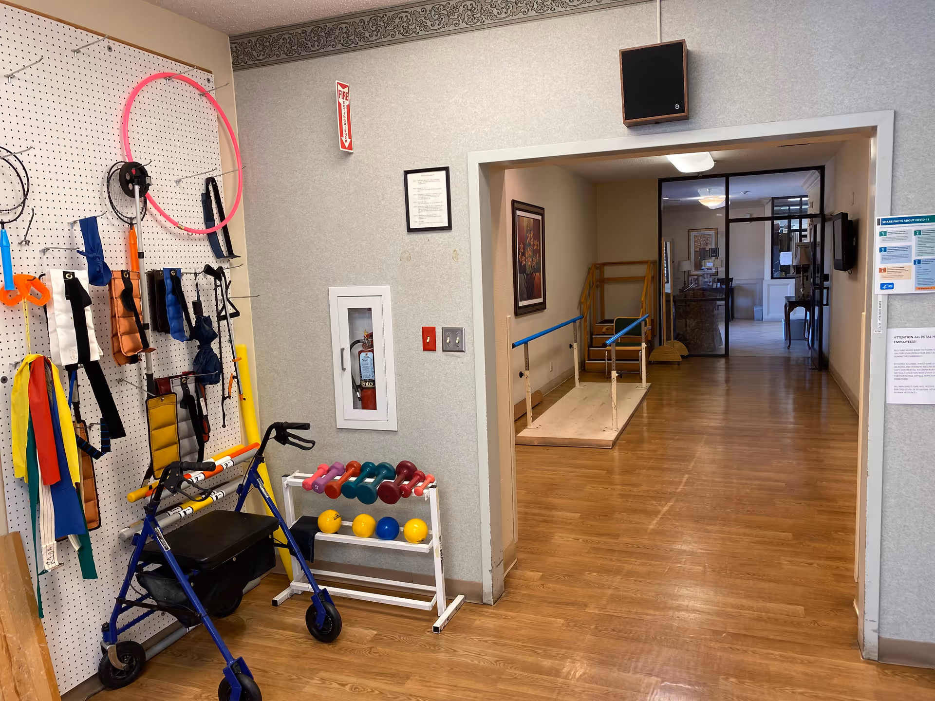 Indoor therapy area with a rollator, colorful hand weights and resistance bands on a pegboard, and parallel bars in a hallway.
