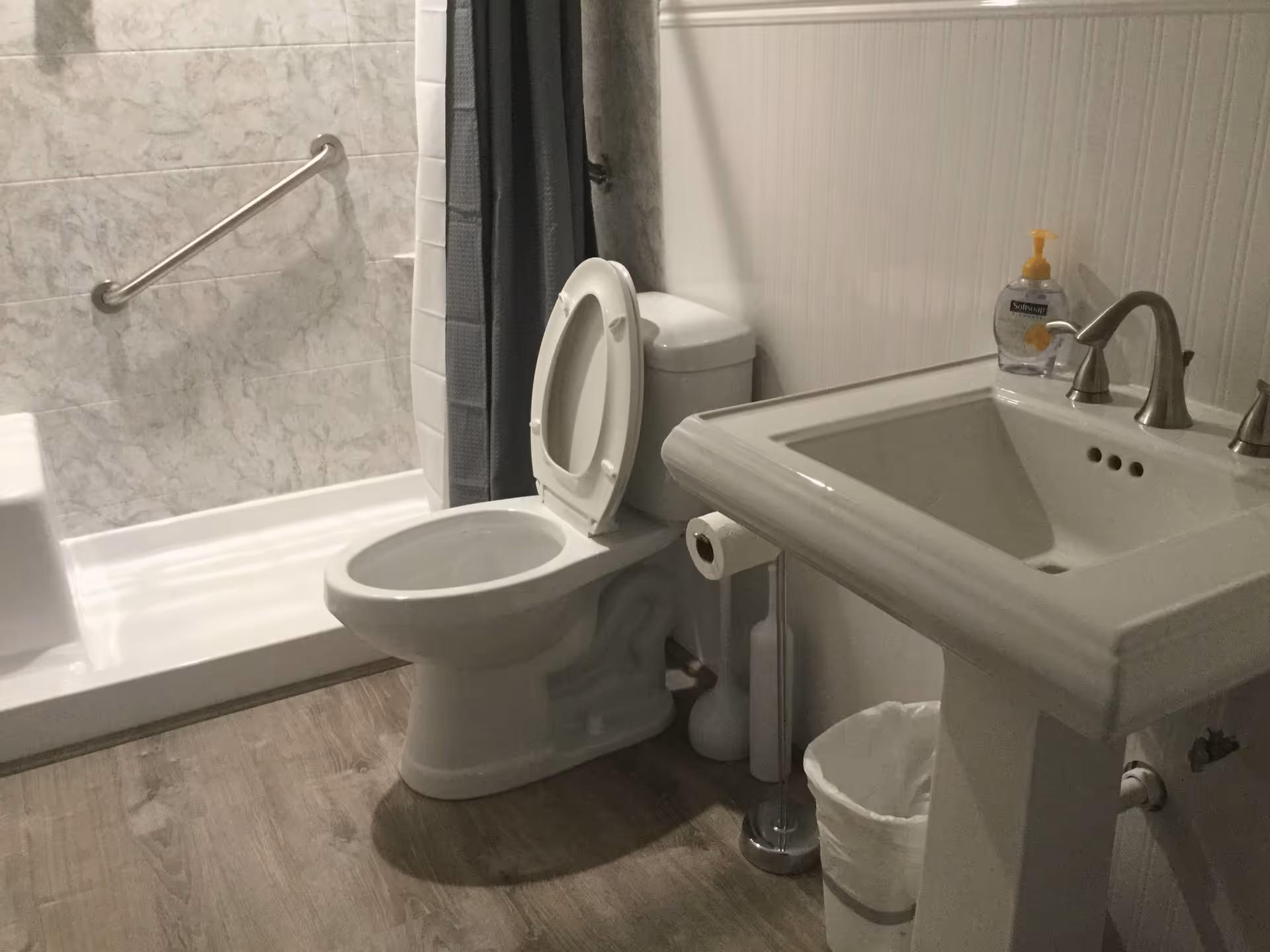A bathroom with a white toilet with the lid open, a white pedestal sink with a soap dispenser on it, a toilet paper holder with a roll of toilet paper, a trash can, and a shower area with a grab bar and a gray shower curtain.