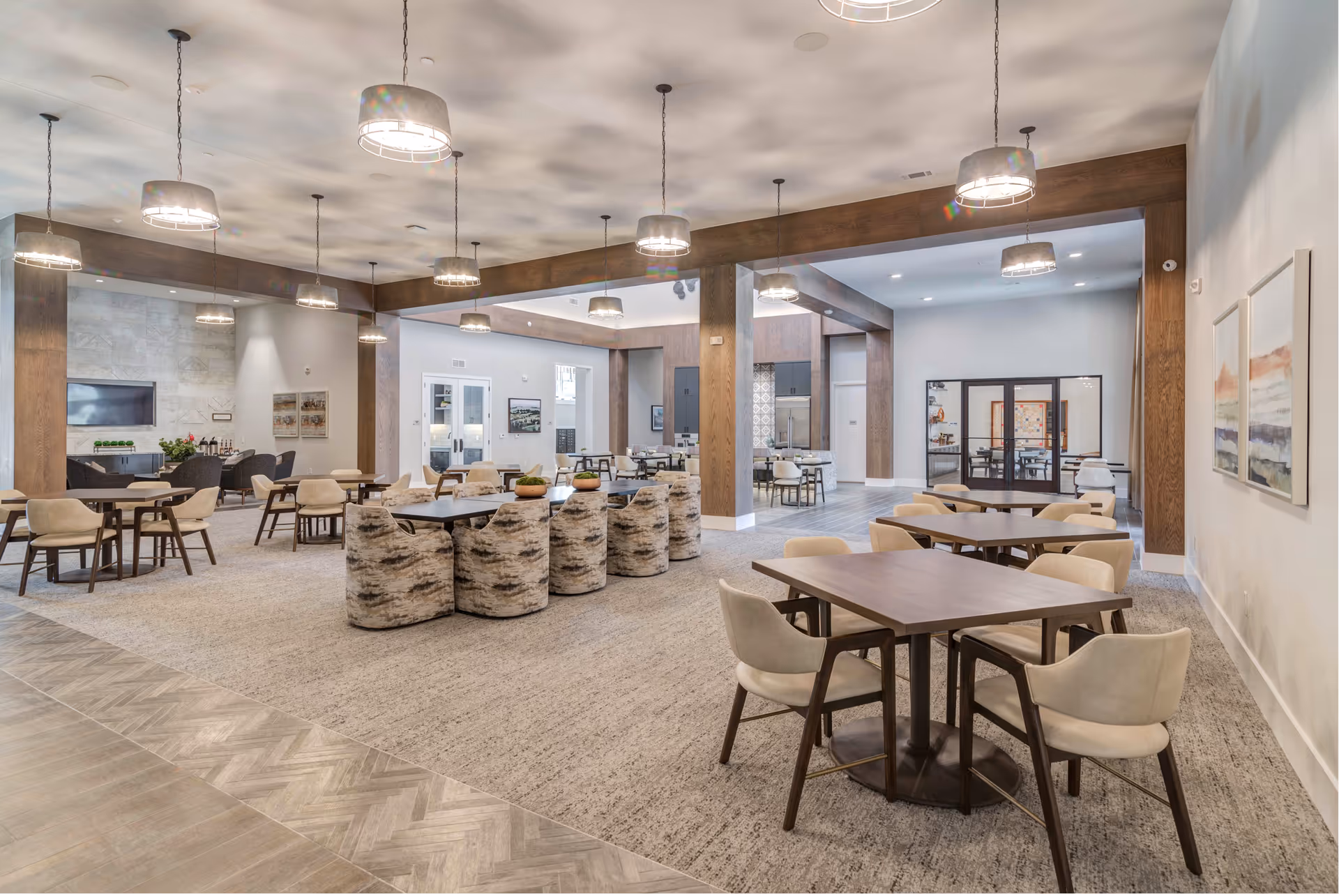 Spacious and modern common area with multiple seating arrangements including tables with chairs and cushioned armchairs. The room features wooden beams, hanging pendant lights, artwork on the walls, and a television mounted on a stone accent wall. The flooring is a combination of carpet and tile.