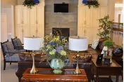 A cozy living room in a retirement community featuring a stone fireplace with a mounted TV above it, flanked by built-in cream-colored cabinets. In front of the fireplace is a brown leather sofa with two brass table lamps on a wooden table, which also holds a green vase with a floral arrangement. Additional seating includes cushioned chairs and a small side table with a plant, creating a warm and inviting atmosphere.