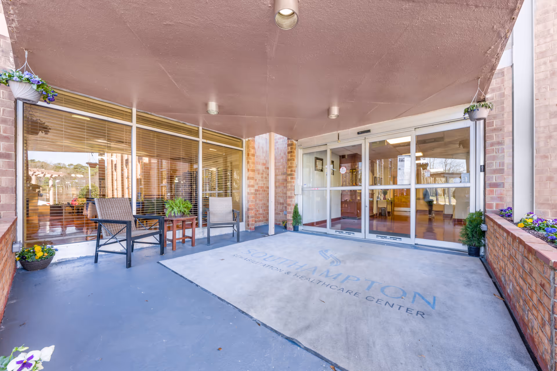 Entrance area of Southampton Rehabilitation & Healthcare Center with a covered porch, two chairs and a small table with a plant, large windows with blinds, and automatic sliding glass doors leading inside. There are flower pots on the sides and the facility name is visible on the floor mat.