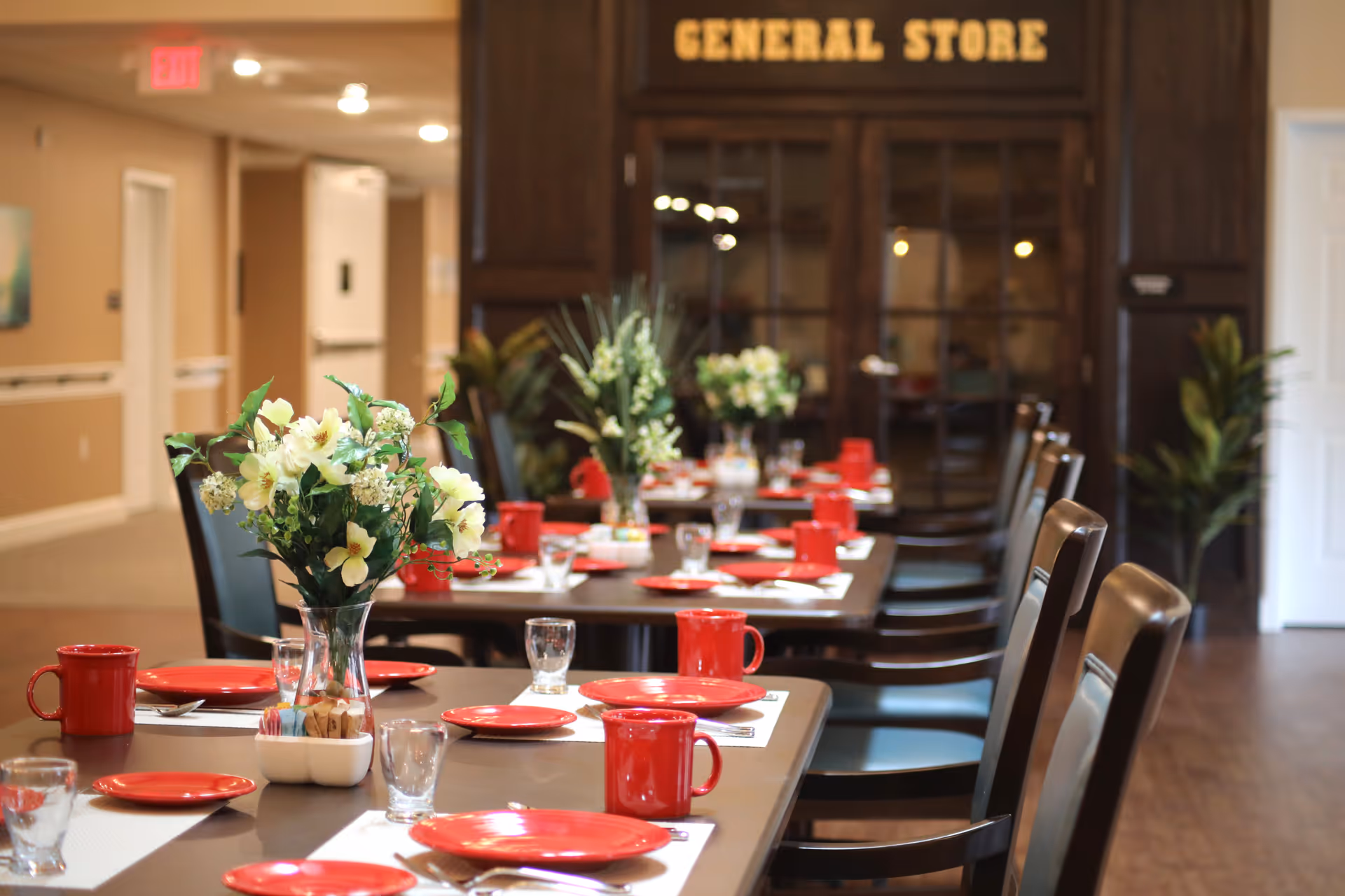 A dining room with a long table set with red plates, red mugs, clear glasses, and white placemats. There are floral centerpieces in glass vases along the table. In the background, there is a dark wooden cabinet labeled 'GENERAL STORE' and some plants near the walls.