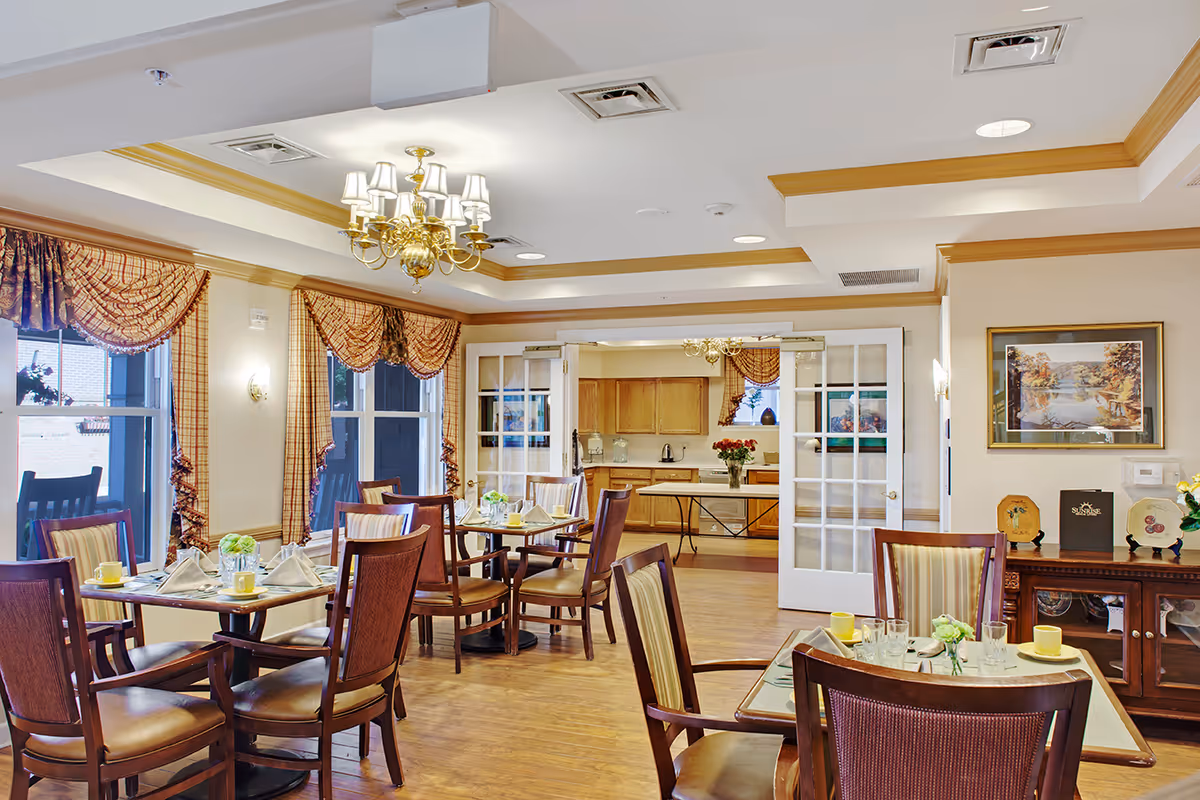 A bright and elegant dining room in a senior living facility with wooden tables and chairs set with plates, glasses, and napkins. The room features large windows with patterned curtains, a chandelier, and a sideboard with decorative items. In the background, there is a kitchen area visible through glass-paneled double doors.