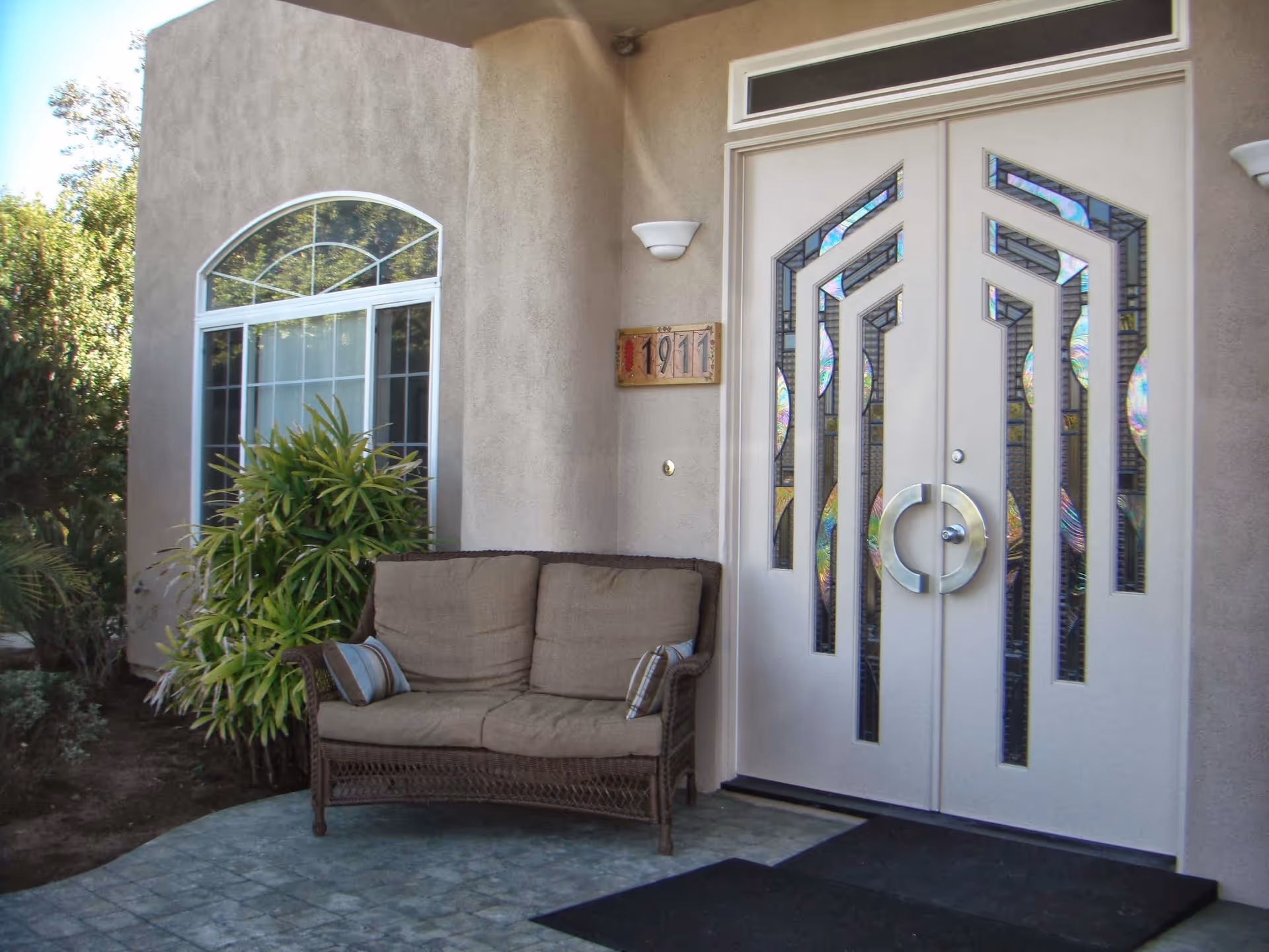 Front entrance of a building with double doors featuring decorative glass panels, a cushioned wicker loveseat with two pillows, a window with an arched top, and some green plants nearby. The building number 1911 is displayed on a plaque next to the door.