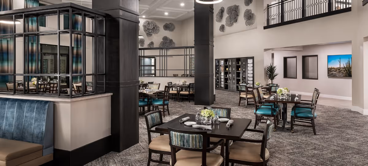 A spacious and elegant dining room with multiple tables and chairs arranged neatly. The room features a high ceiling with modern light fixtures, decorative wall art, and a mix of upholstered and wooden chairs. There is a glass partition on the left side and a carpeted floor throughout the area.