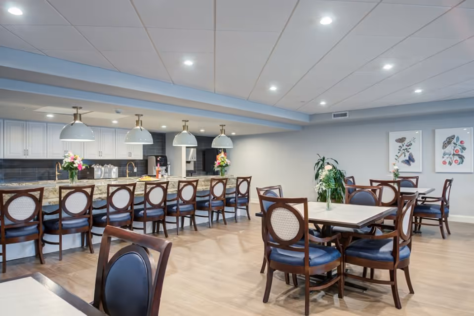 A bright and spacious dining area with several tables and chairs arranged neatly. There is a long counter with high chairs and pendant lights hanging above it. The background features a kitchen area with white cabinets and a dark backsplash. The room is decorated with flower vases on the tables and framed botanical artwork on the walls.