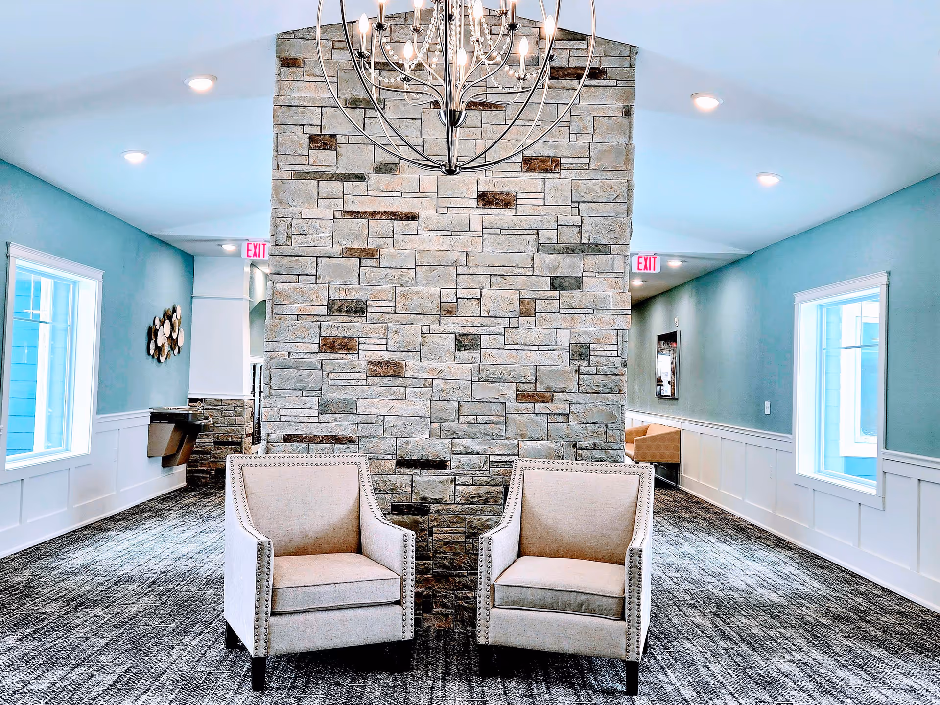 Two beige armchairs sit in front of a stone partition in a bright communal lounge with teal walls and a chandelier.