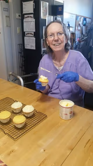 An elderly woman wearing a purple top and blue gloves is sitting at a wooden table, smiling while frosting a cupcake. Several unfrosted cupcakes are on a cooling rack on the table, along with a container of frosting and a spatula. The background shows a kitchen area with a refrigerator and bulletin boards.