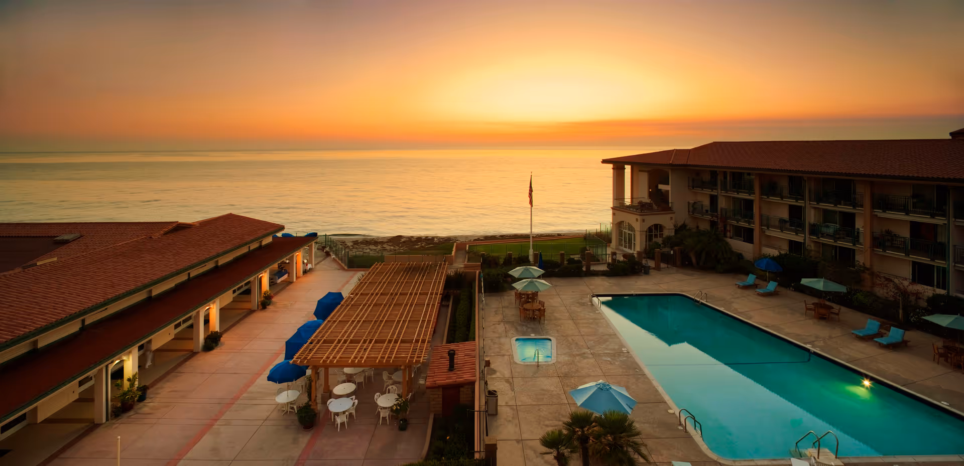 Outdoor view of a senior living facility at sunset featuring a swimming pool with lounge chairs and umbrellas, adjacent buildings with balconies, and the ocean in the background under a colorful sky.