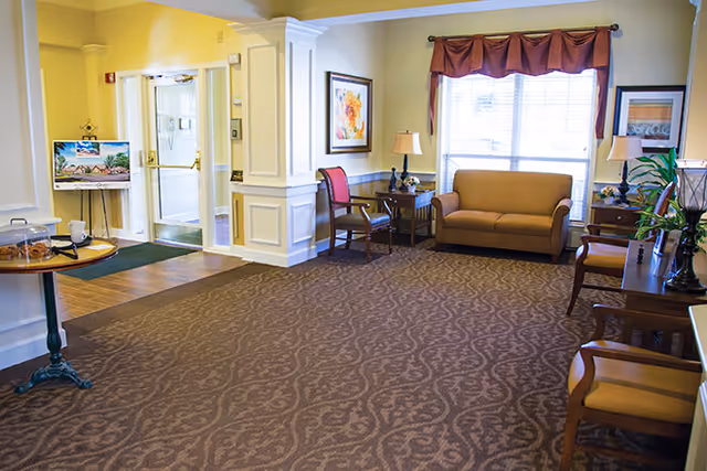 A well-lit living room area with patterned carpet, a brown sofa, several wooden chairs with cushions, side tables with lamps, framed artwork on the walls, and a large window with a valance curtain letting in natural light. There is also a small round table with a glass container and a painting on an easel near the entrance.