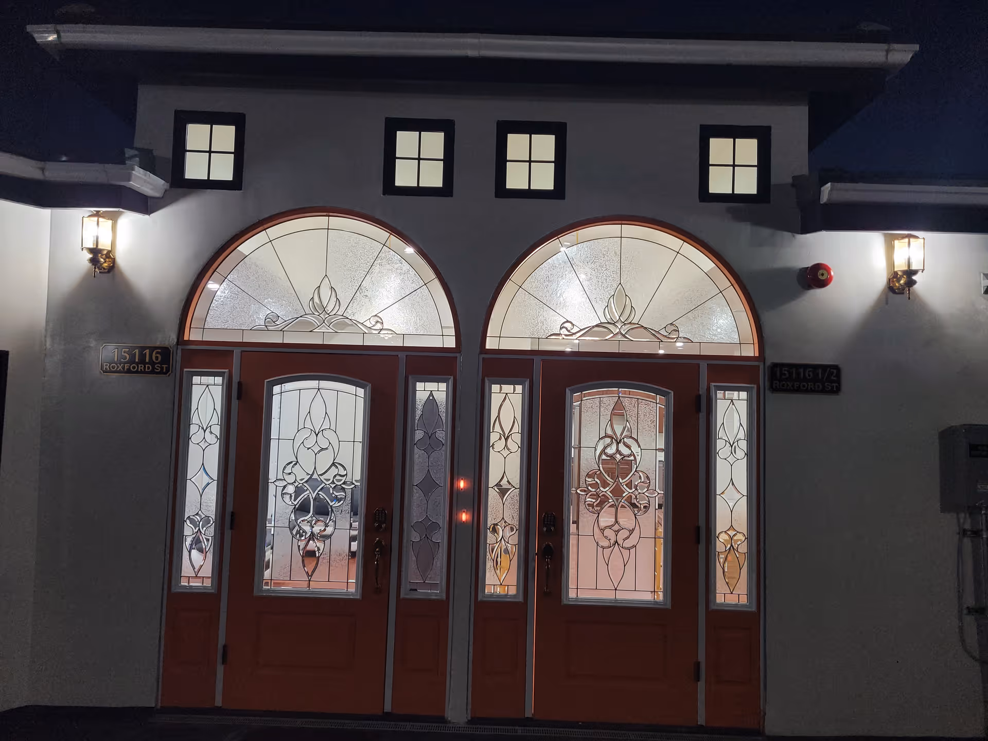 Ornate double front doors with arched stained-glass transoms and two illuminated wall lanterns at night.