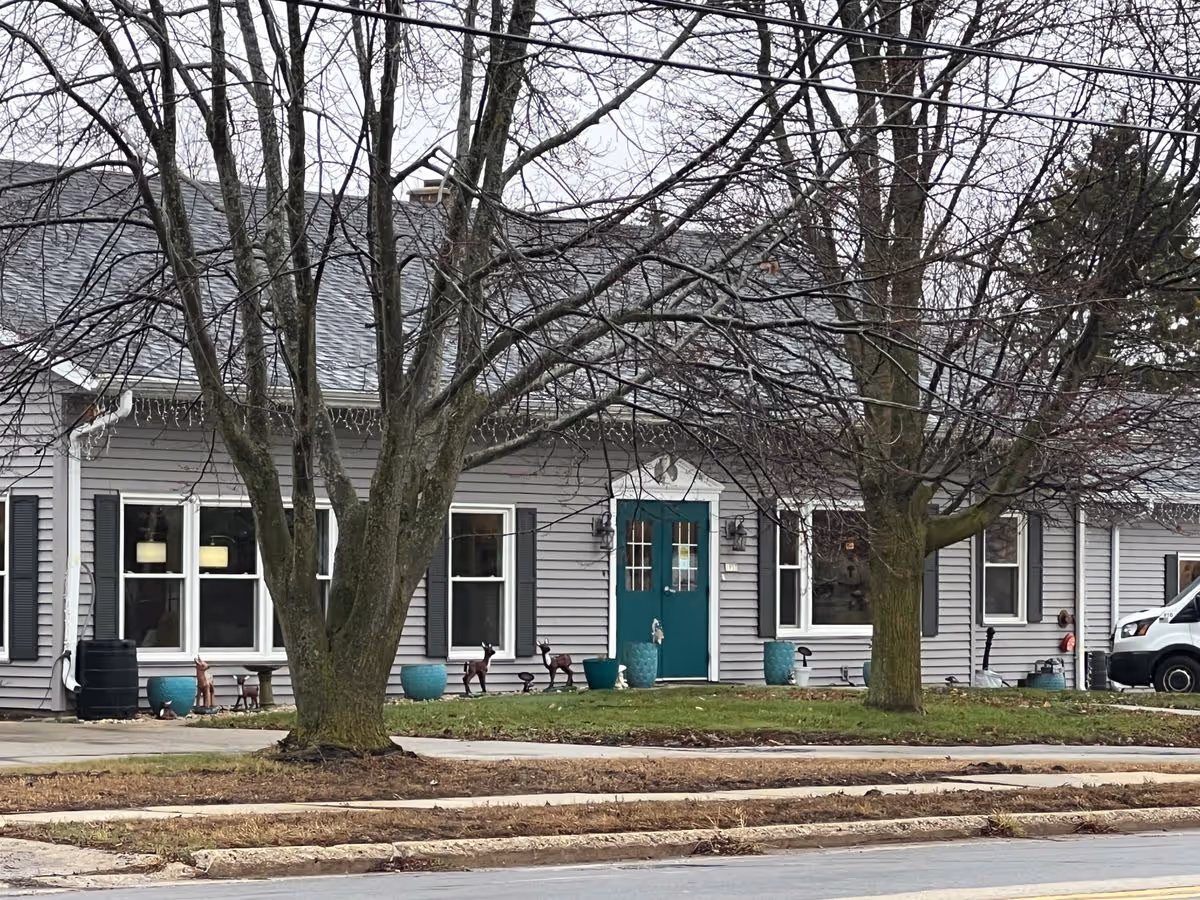 Front exterior of a single-story gray building with a teal front door, two leafless trees, and potted plants on the lawn.