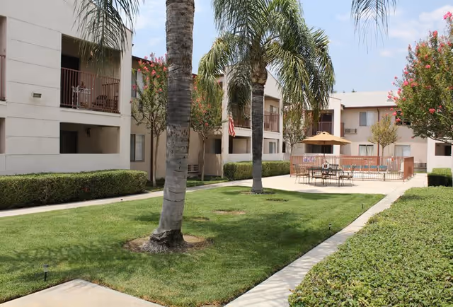 Outdoor courtyard area of Parkside Senior Apartments featuring green grass, palm trees, trimmed hedges, and a seating area with tables and chairs under a large umbrella. The courtyard is surrounded by a two-story apartment building with balconies and windows.