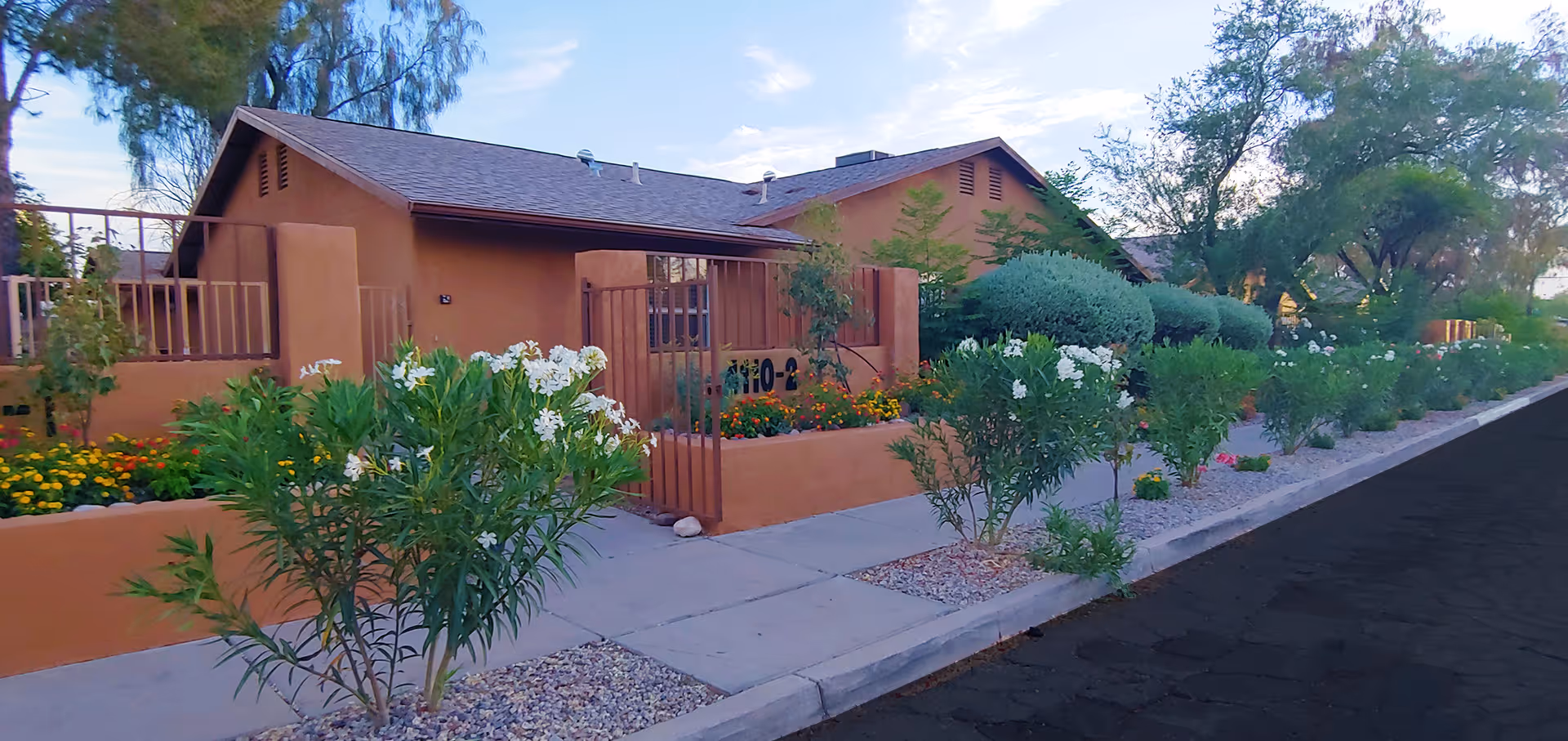 Single-story tan building with a gated entrance and landscaped front yard along a sidewalk.