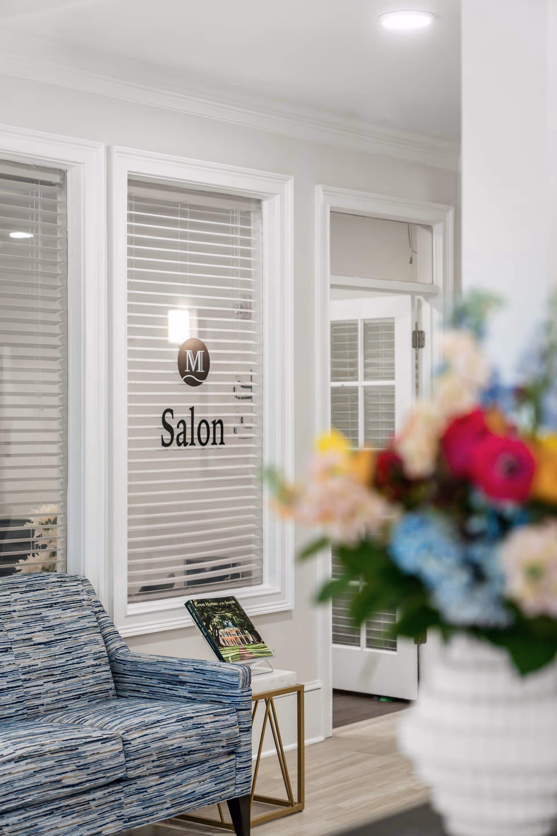 Interior view of a senior living facility showing a blue patterned armchair next to a small table with a book on it. Behind the chair is a window with blinds and a sign that reads 'Salon'. A door with glass panels is visible in the background. A blurred bouquet of colorful flowers is in the foreground on the right side.