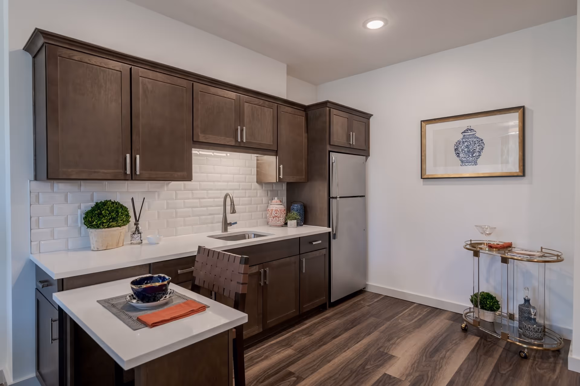 A modern kitchen with dark wood cabinets, white countertops, and a white subway tile backsplash. There is a stainless steel refrigerator, a small sink, and a countertop with a place setting including a bowl, plate, and orange napkin. On the right side, there is a gold bar cart with decorative items and a framed picture of a blue and white vase hanging on the wall. The floor is wood with a dark finish.