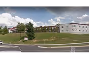 Exterior view of a multi-story nursing home building situated on a grassy hill with a few trees and a road in the foreground under a partly cloudy sky.