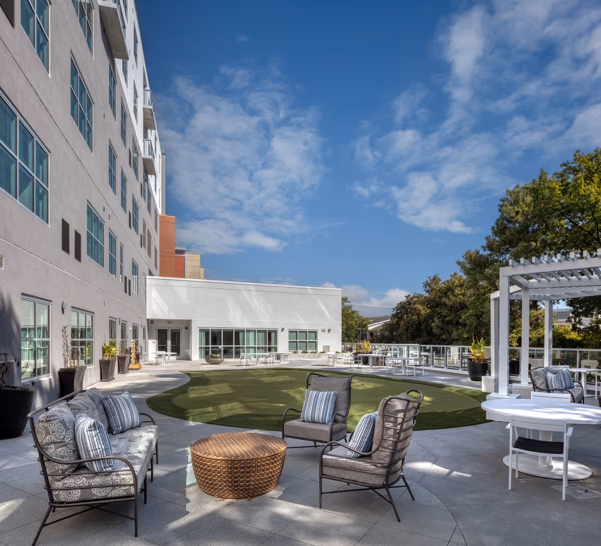 Outdoor patio area at Heartis Buckhead Senior Living featuring cushioned chairs and a sofa arranged around a round wicker table, a green artificial turf lawn, white tables and chairs, potted plants, and a pergola under a partly cloudy blue sky.
