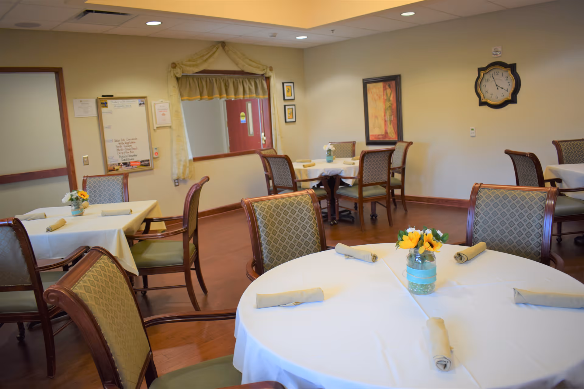A dining room with several tables covered in white tablecloths and set with rolled napkins. Each table has a small vase with yellow flowers. The room has wooden chairs with patterned upholstery, a wall clock, framed artwork, and a window with a curtain. The floor is wooden and the walls are painted beige.