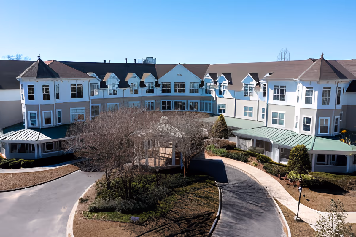 Front exterior view of a large, multi-story senior living facility building with white walls, multiple windows, and a brown roof. The building is surrounded by landscaped grounds with trees, bushes, and a curved driveway leading to the entrance under a green awning. The sky is clear and blue.