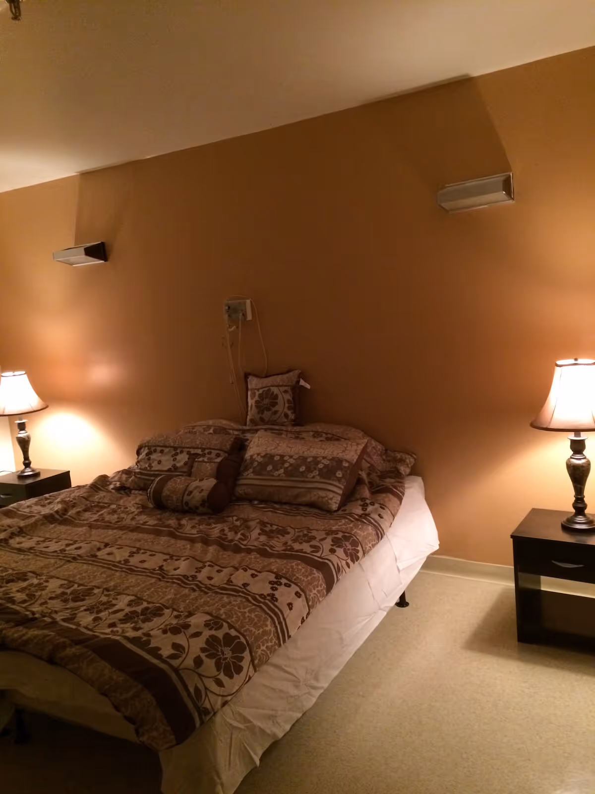 A bedroom with a bed covered in a brown floral patterned comforter and multiple pillows. There are two bedside tables, each with a lamp emitting warm light. The walls are painted a warm beige color, and there are two wall-mounted speakers above the bed.