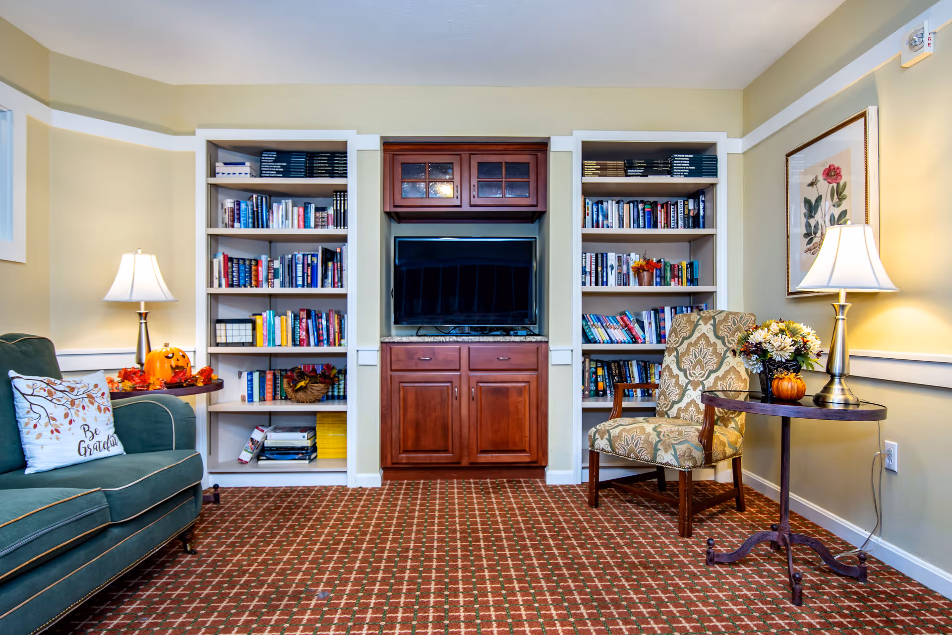 Comfortable lounge area with bookshelves and a central TV cabinet, upholstered sofa and chair, side tables with lamps and seasonal decorations.