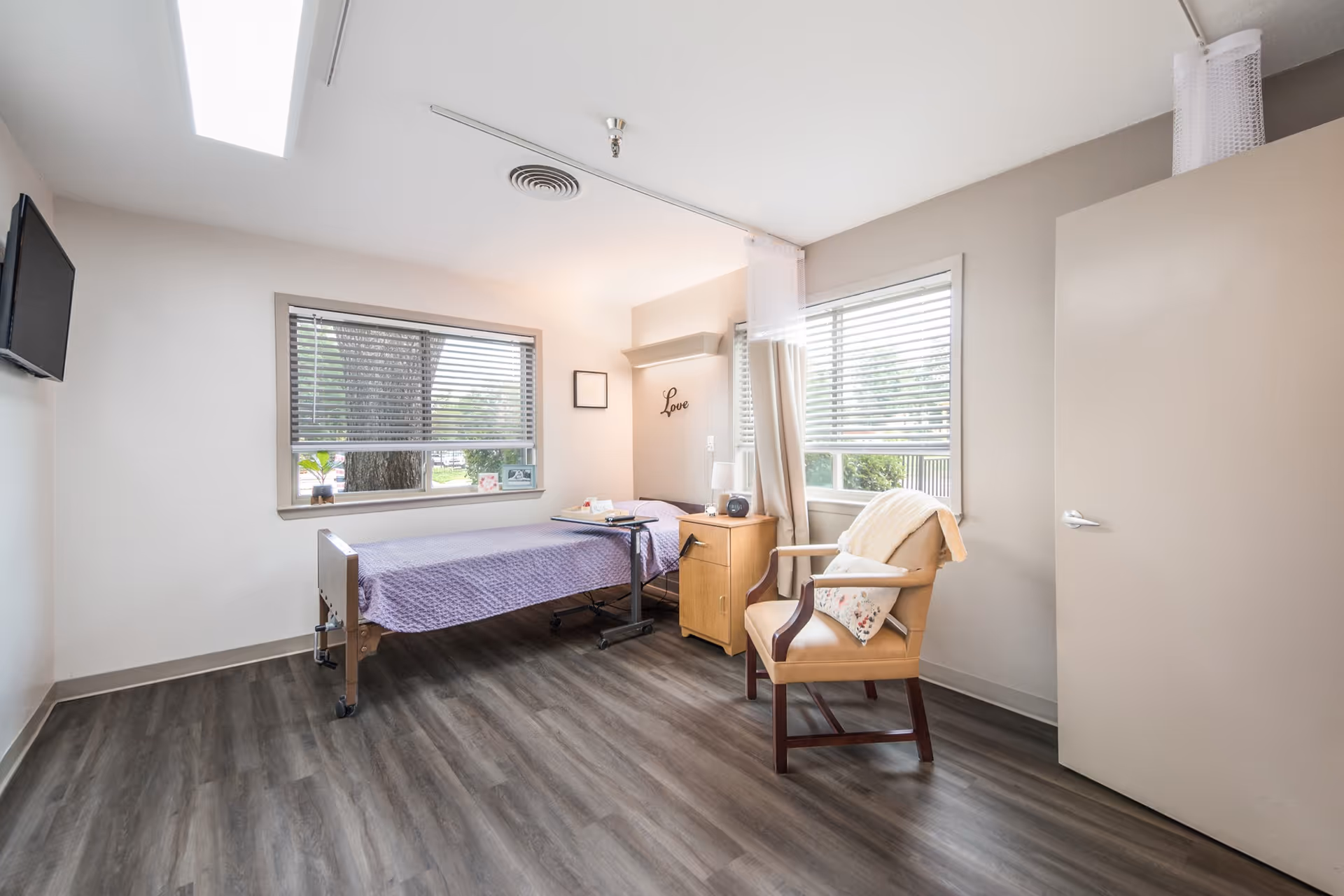 Bright senior care bedroom with a single medical bed, bedside table, armchair and two windows.