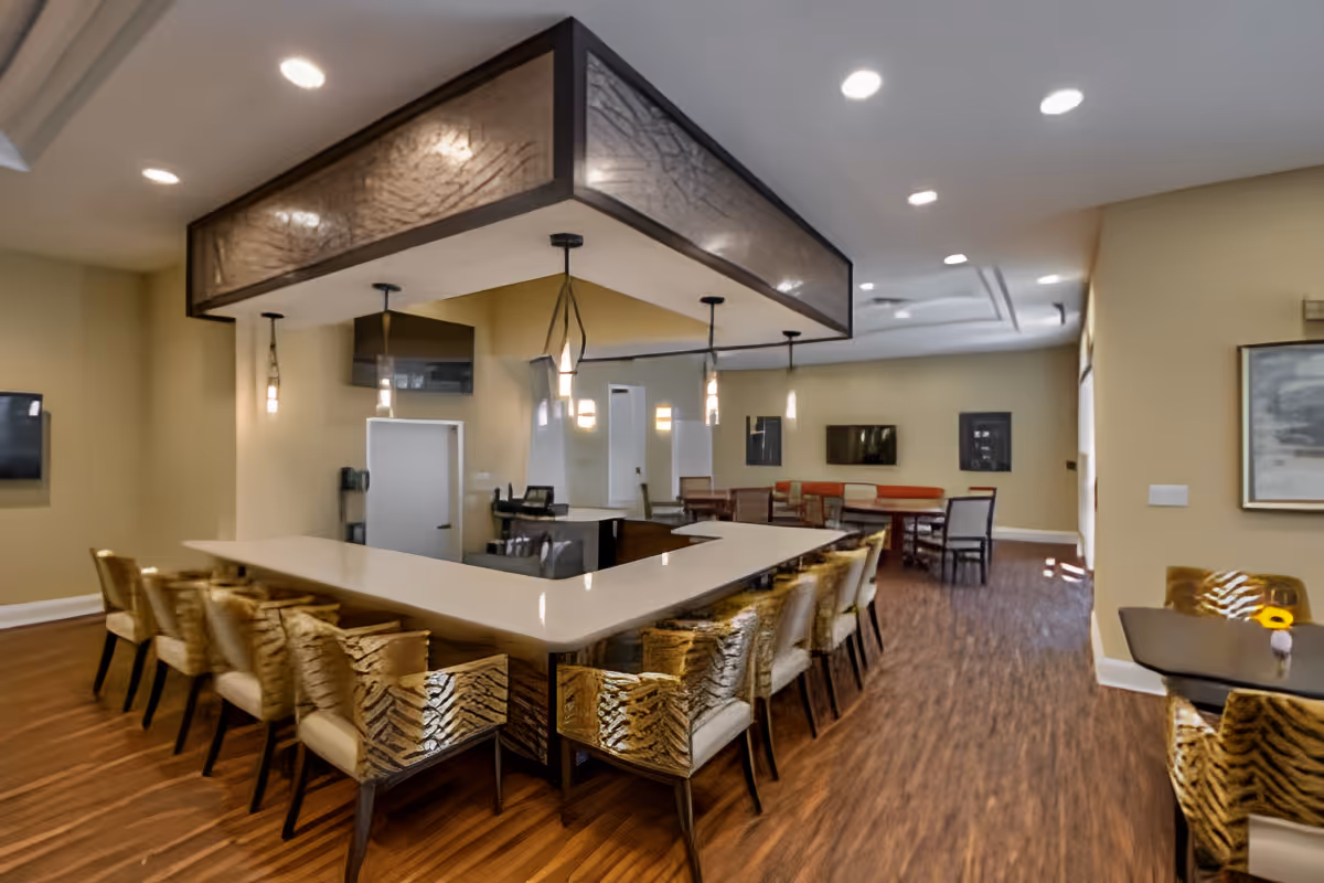 Interior view of a senior living facility lounge area featuring a large U-shaped counter with multiple patterned chairs around it. The room has warm beige walls, wooden flooring, pendant lights hanging above the counter, and additional seating with tables and chairs in the background. There are two wall-mounted televisions and framed artwork on the walls.