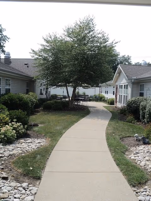 A curved concrete pathway runs between two single-story buildings with beige siding. The path is bordered by small landscaped areas with grass, bushes, and decorative rocks. A large tree stands near the center of the image, with outdoor tables and chairs visible in the background under the tree. The sky is bright and clear.