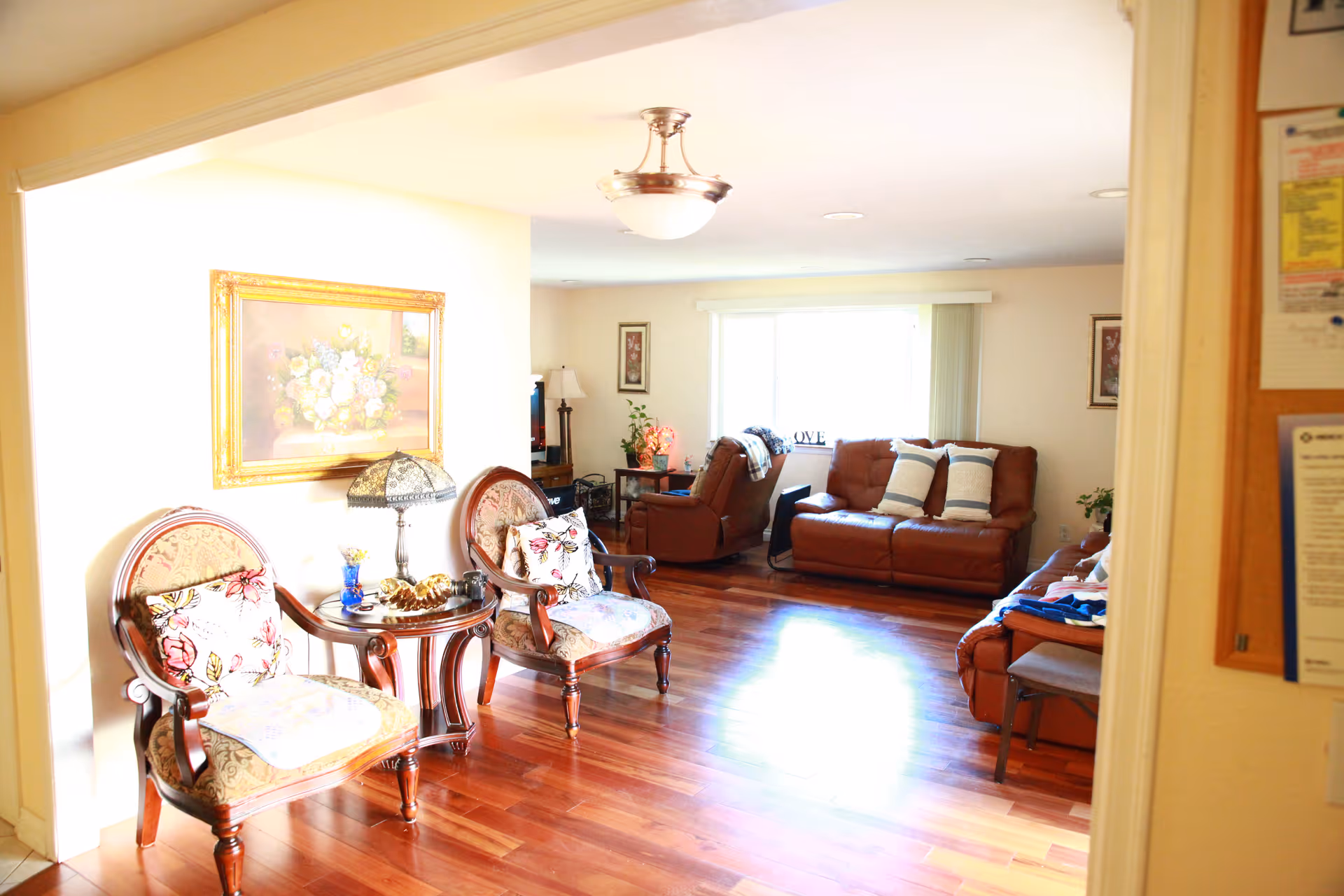 Bright living room with hardwood floors, two upholstered armchairs and side table in the foreground and leather sofas by a large window in the background.