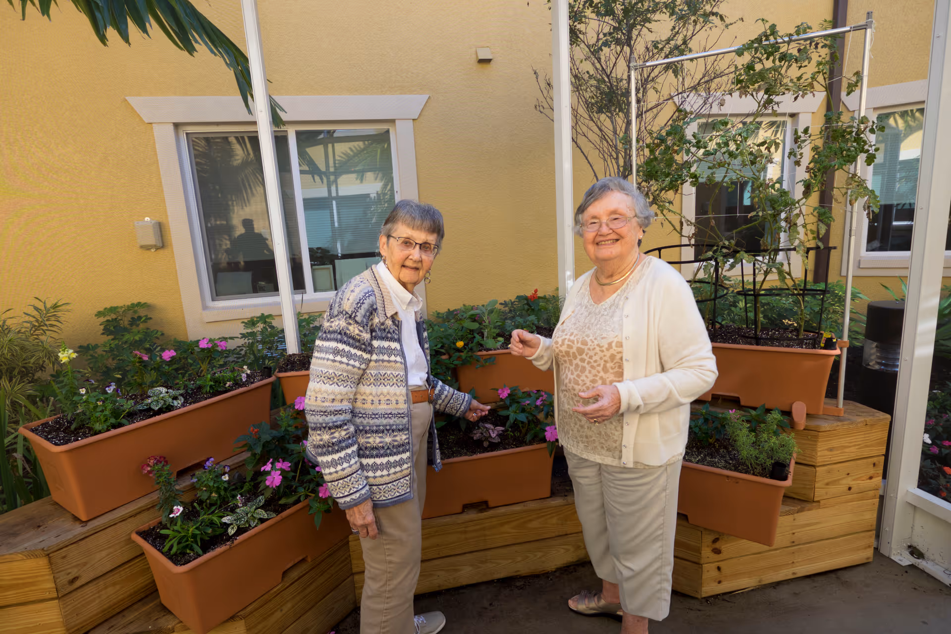 Two elderly women standing and smiling in an outdoor garden area with multiple planter boxes filled with various flowers and plants, against a yellow building wall with windows.