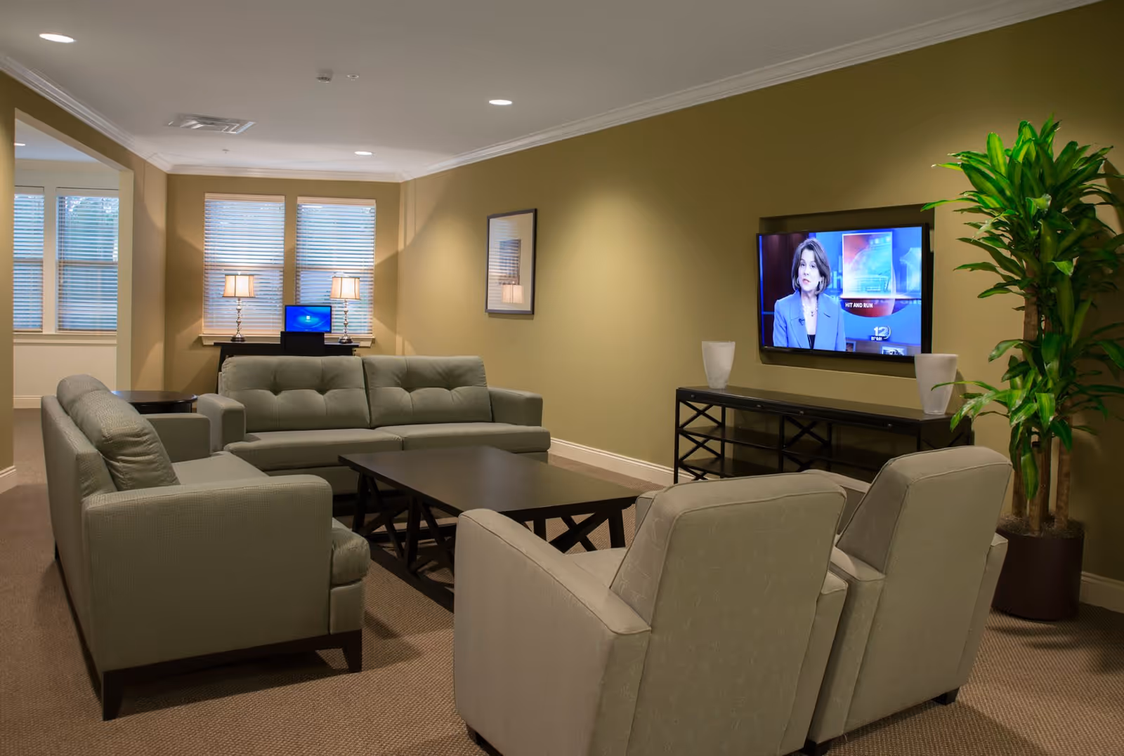 A cozy living room with beige walls and carpet, featuring a green sofa, two matching armchairs, a dark wooden coffee table, a wall-mounted flat-screen TV showing a news anchor, a black console table with two white vases, a large potted plant, and a desk with a laptop and two lamps near three windows with blinds.