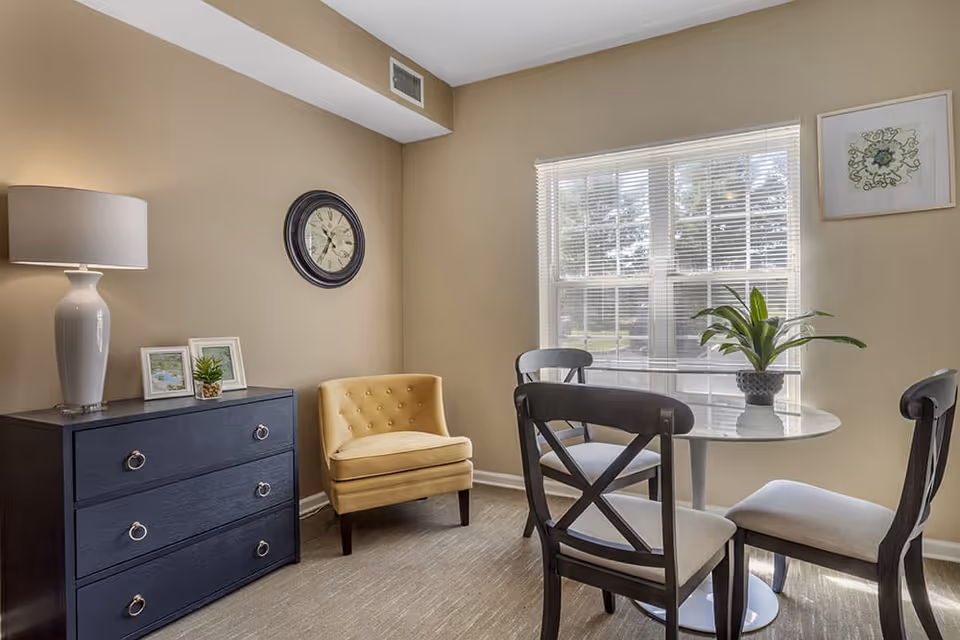 A cozy interior room with beige walls featuring a small round dining table with four chairs, a yellow upholstered armchair, a dark blue chest of drawers with a white lamp and framed pictures on top, a wall clock, and a window with white blinds letting in natural light. A potted plant is placed on the dining table.