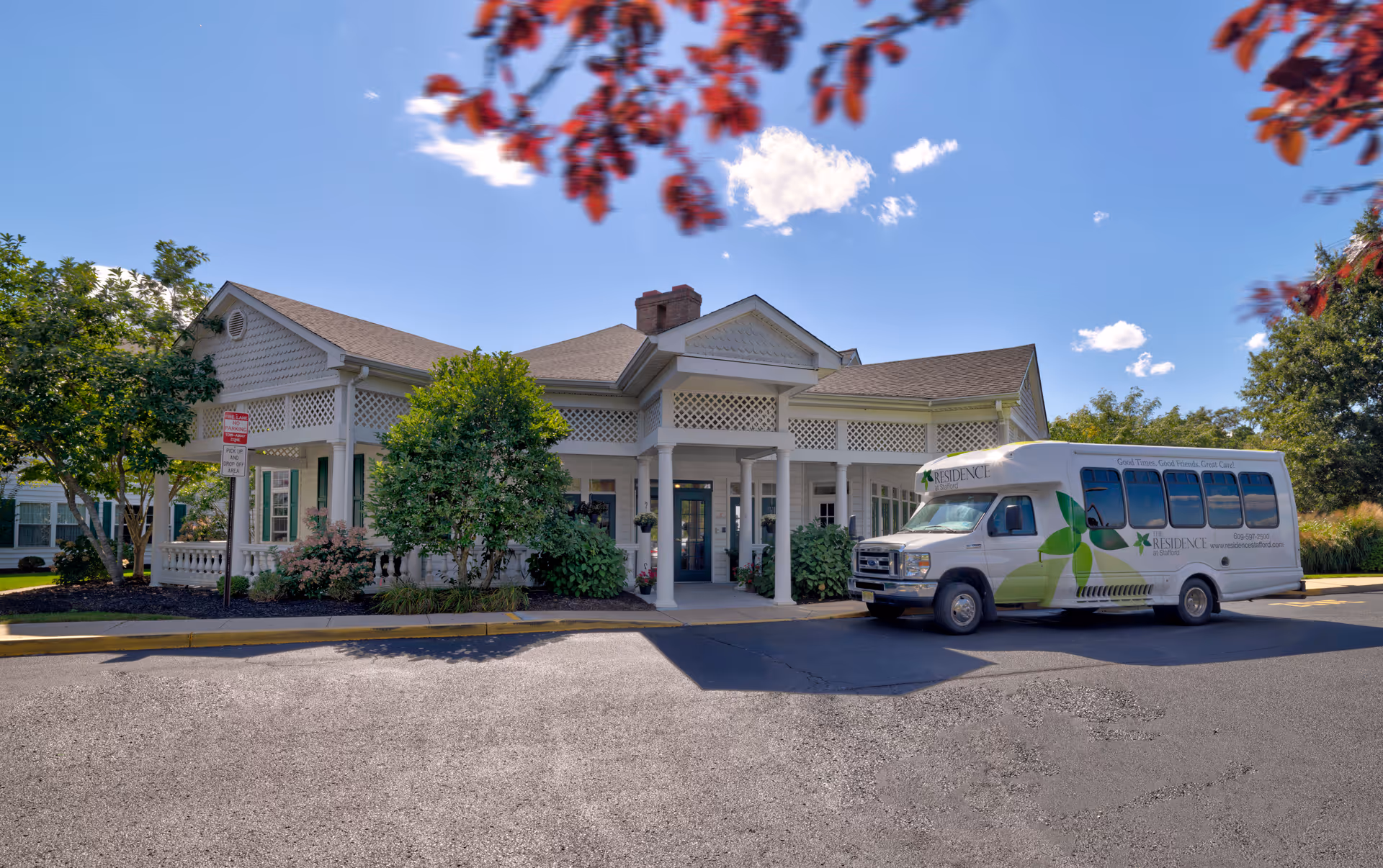 Exterior view of The Residence at Stafford building with a covered entrance, surrounded by trees and bushes under a clear blue sky. A white shuttle bus with green leaf graphics and the facility's name is parked in front.