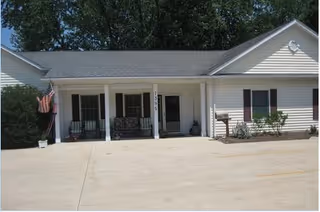 Front exterior view of a single-story building with white siding, a gray shingled roof, and a covered porch area with seating. There is an American flag on the left side near some bushes and a mailbox on the right side near some plants.