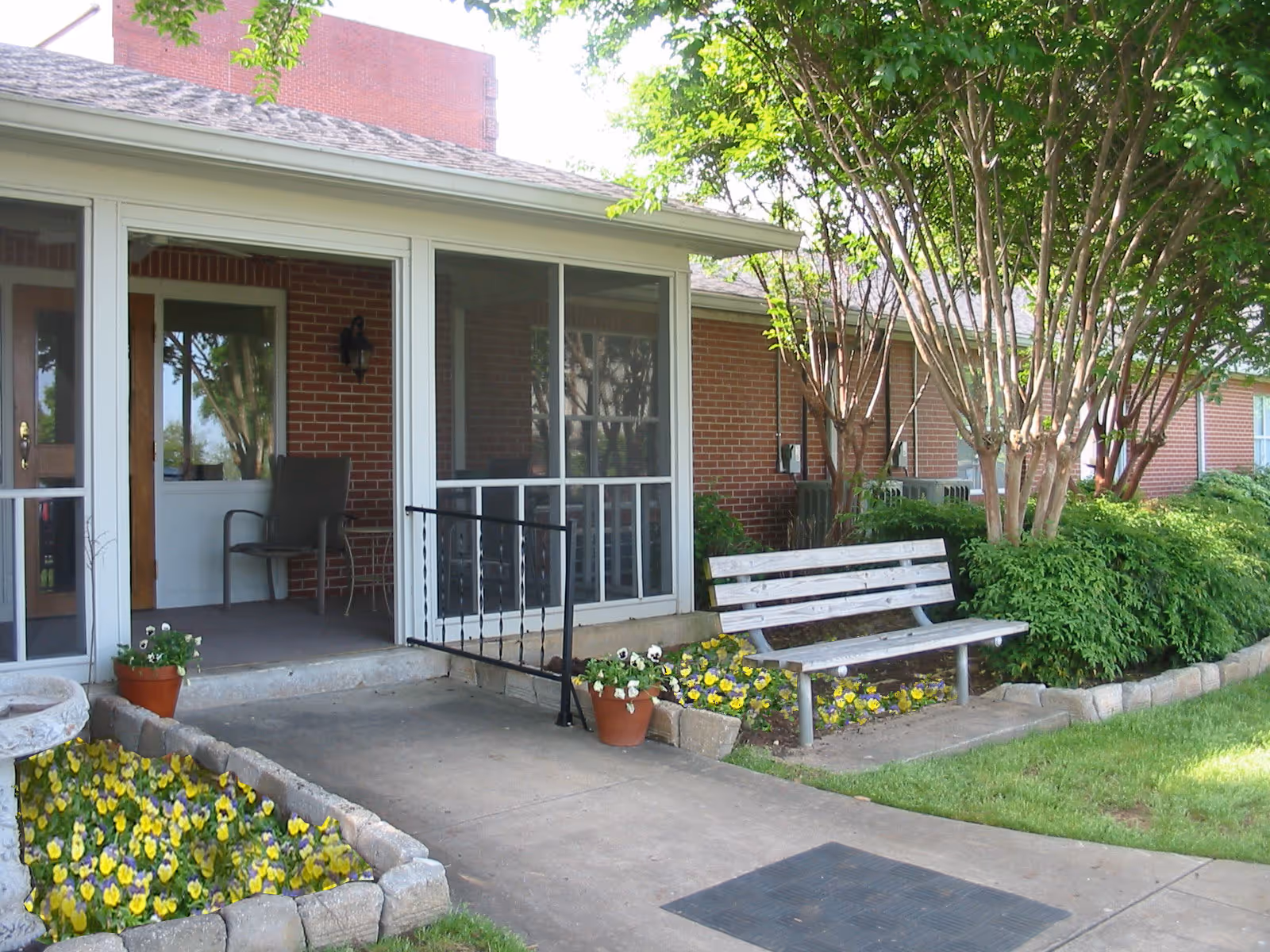 Outdoor view of a senior living facility entrance with a screened porch, a wooden bench, flower beds with yellow and purple flowers, and green shrubs and trees surrounding the area.