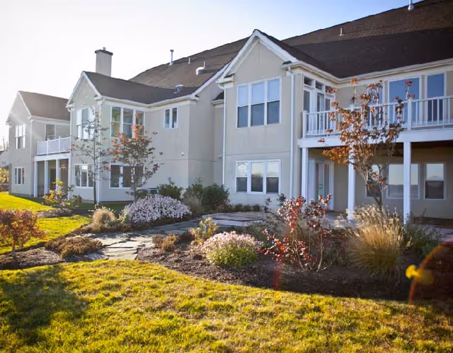 Exterior view of a senior living facility building with beige walls and a brown roof, featuring multiple windows and balconies. The foreground shows a landscaped garden with grass, shrubs, and small trees under bright sunlight.