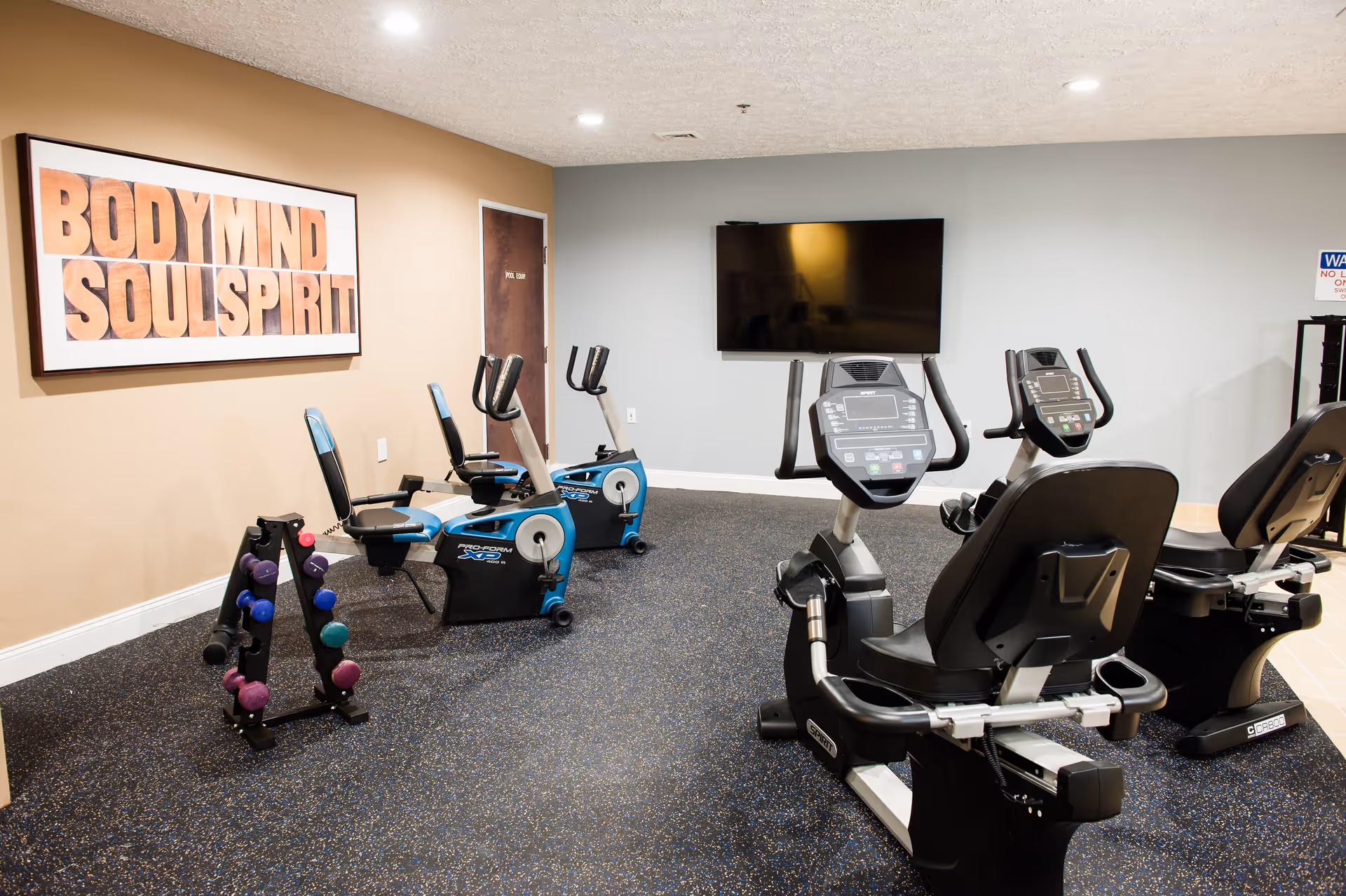 A senior living facility exercise room with stationary recumbent bikes, a rack of colorful dumbbells, and a large wall-mounted TV. A framed sign on the wall reads 'BODY MIND SOUL SPIRIT'.