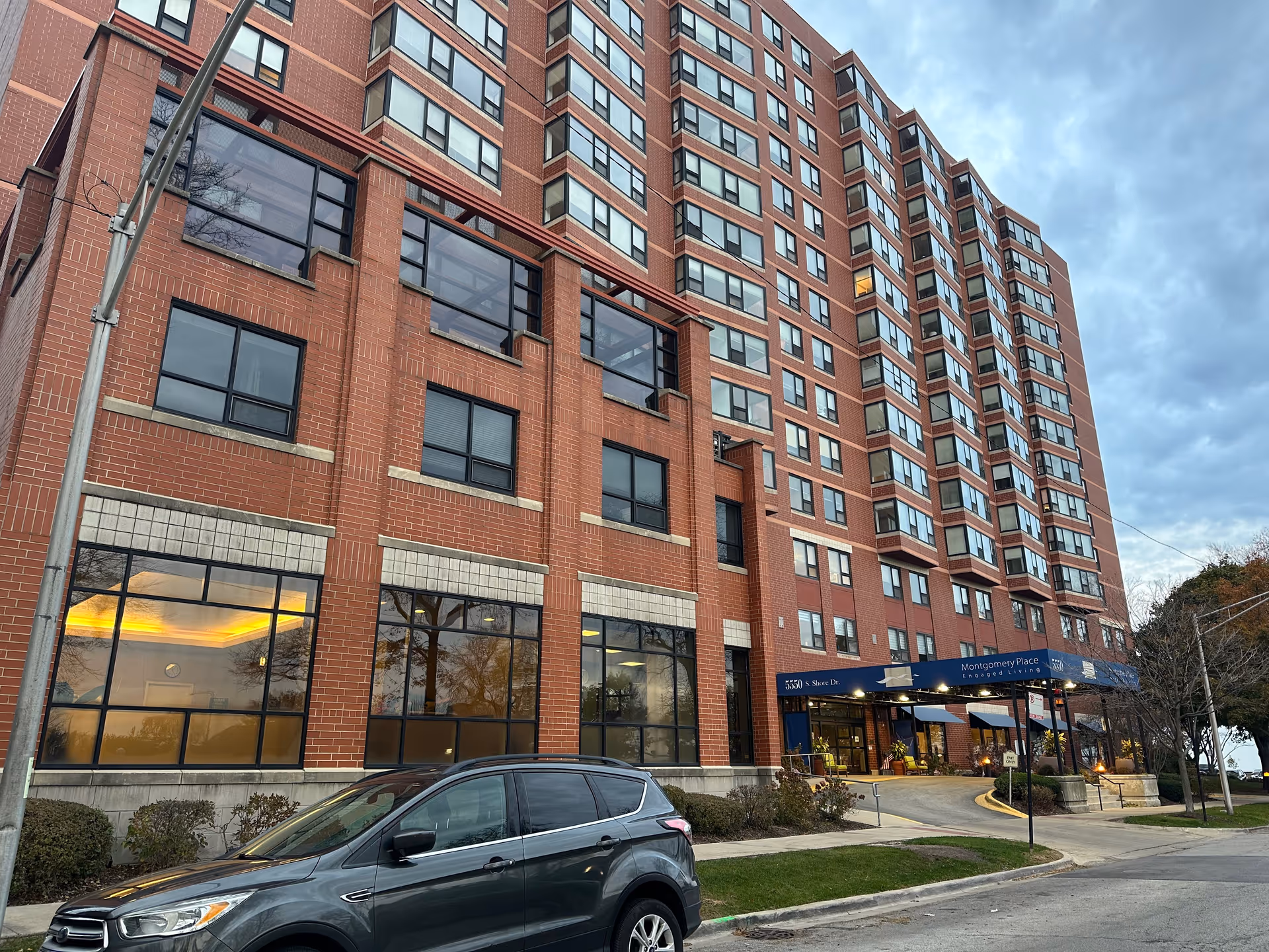 Exterior view of a multi-story brick building with many windows, identified as Montgomery Place Retirement Community. A dark-colored SUV is parked in front of the building near the entrance, which has a blue awning with the facility's name. The sky is cloudy and it appears to be late afternoon or early evening.