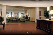 Interior view of a senior living facility lobby or common area with a reception desk on the right, seating area with chairs and tables in the background, potted plants, and framed artwork on the walls.