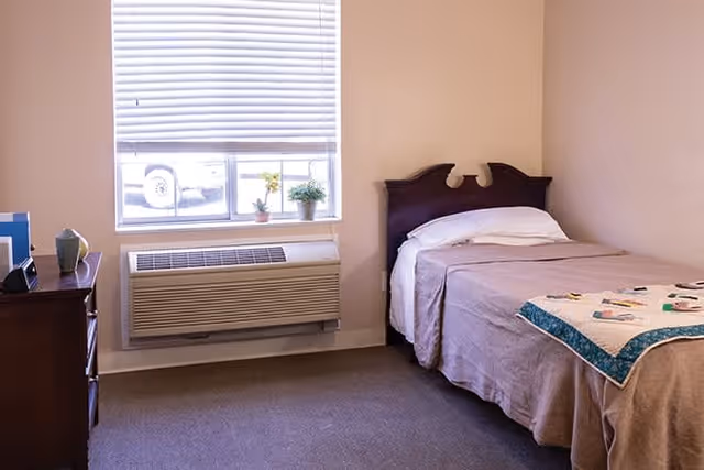 A simple bedroom with a single bed covered with a beige blanket and a colorful quilt at the foot. There is a wooden headboard and a pillow on the bed. Next to the bed is a window with closed blinds, and below the window is an air conditioning unit. On the left side of the image, there is a wooden dresser with a few decorative items on top. The walls are painted light beige and the floor is carpeted.