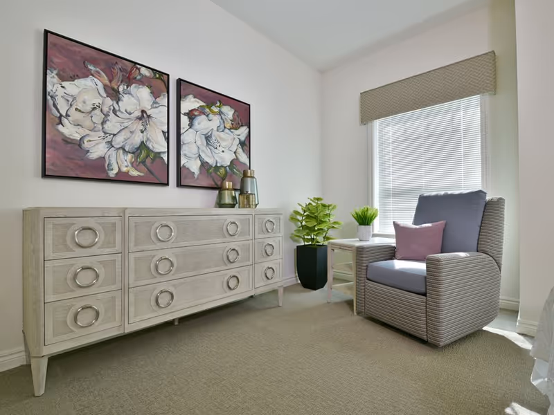 A bright room featuring a striped armchair by a window, a light-wood dresser with circular handles, potted plants, and two floral paintings on the wall.