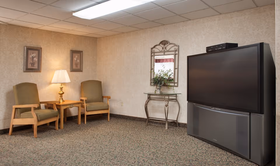 Small sitting area with two armchairs and a side table with a lamp, decorative mirror and a large television in a carpeted room.