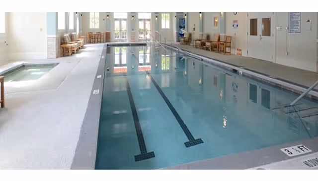 Indoor lap swimming pool with lane markings, chairs along the deck, and a small hot tub at one end.