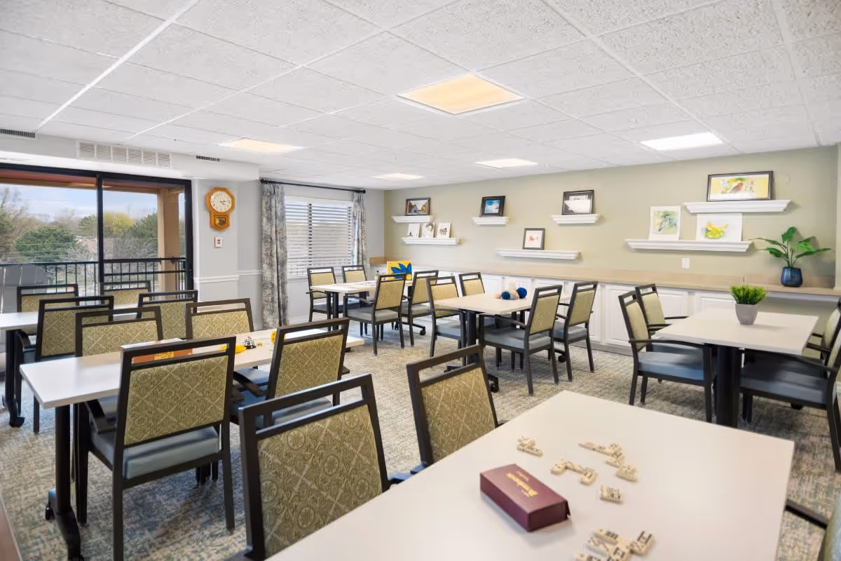 A bright and spacious dining room with multiple tables and chairs arranged neatly. The room has large windows with curtains allowing natural light to enter, and shelves on the wall displaying framed pictures and small plants. There is a clock on the wall near the window and some game pieces on the tables.