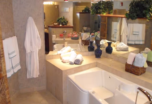 A bathroom with a large bathtub featuring a built-in seat, beige tiled walls and floor, a countertop with rolled white towels, two dark blue vases, a basket with towels, a white bathrobe hanging on the wall, and a large mirror reflecting the room and plants.